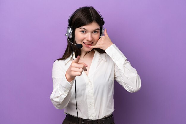 Telemarketer donna russa che lavora con un auricolare isolato su sfondo viola che fa il gesto del telefono e indica la parte anteriore