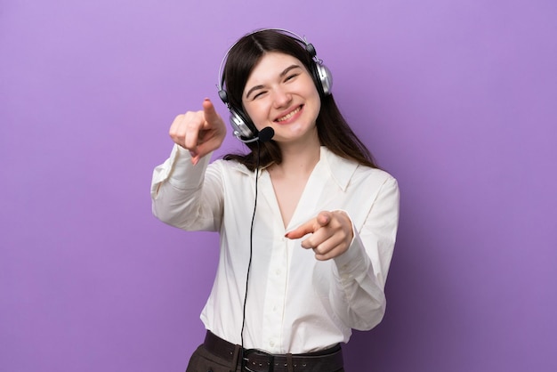 Telemarketer Russian woman working with a headset isolated on purple background points finger at you while smiling