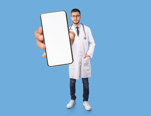 Телемедицинский молодой врач-мужчина в форме показывает большой пустой смартфон в руке