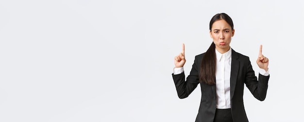 Teleurgesteld pruilende Aziatische vrouwelijke ondernemer in pak klagen en mokken van mislukking wijzende vingers omhoog grimassen ontevreden gevoel neer van spijt staande witte achtergrond