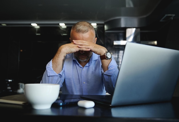 Teleurgestelde zakenman van middelbare leeftijd die aan een tafel zit met een laptop, naar beneden kijkt en zijn gezicht zorgvuldig verbergt met zijn handen tegen de achtergrond van de moderne keuken