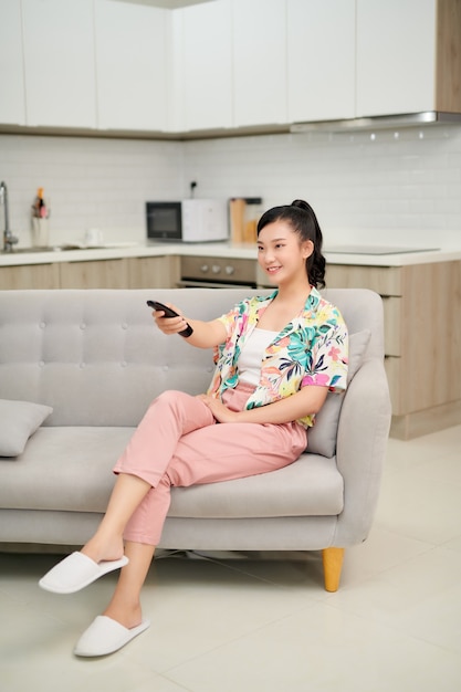 Television, relax, home and happiness concept - smiling teenage girl sitting on couch with tv remote control at home