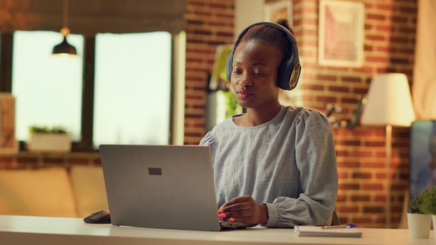 Teleworker listening to music and solving daily tasks, self employed woman. African american girl having fun with headset while she works on duties during sunset hour at home. Handheld shot.