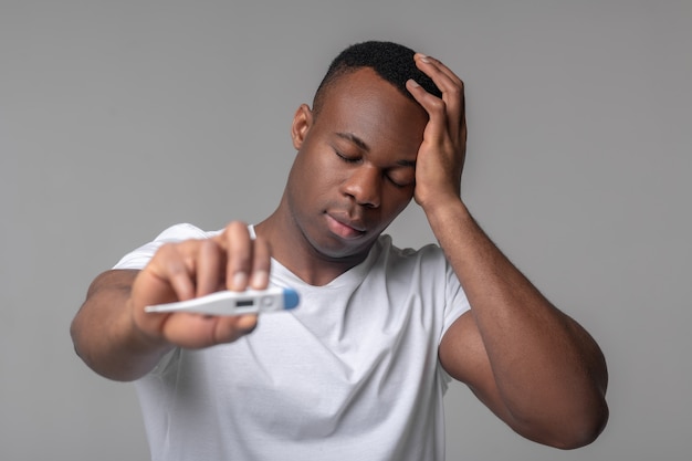 Temperatuur, virus. Jonge aantrekkelijke Afro-Amerikaanse man in witte t-shirt met gesloten ogen weergegeven: thermometer hand aan hoofd aan te raken