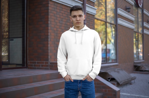 Template male white hoodie on a guy, front view, presentation of clothes on the street. Mockup hoods on the background of a brick building. Place for your pattern and design.
