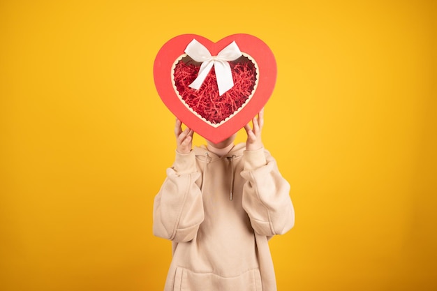 Ten years old boy holding a heartshaped gift box in his hand no faceTeen boy in biege hoodie on yellow background