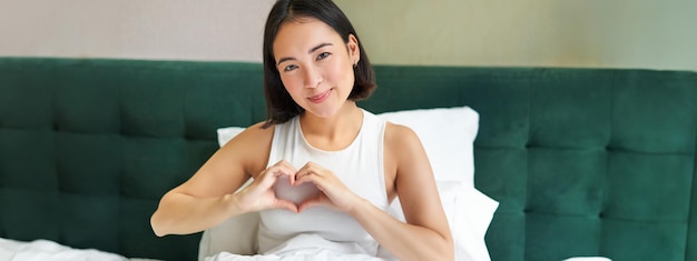 tender asian girl shows heart sign lying in bed spending time in bedroom expresses love and care