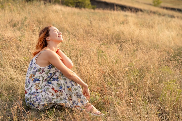 Tender beautiful red-haired girl enjoys the sunset in a field with a hill.