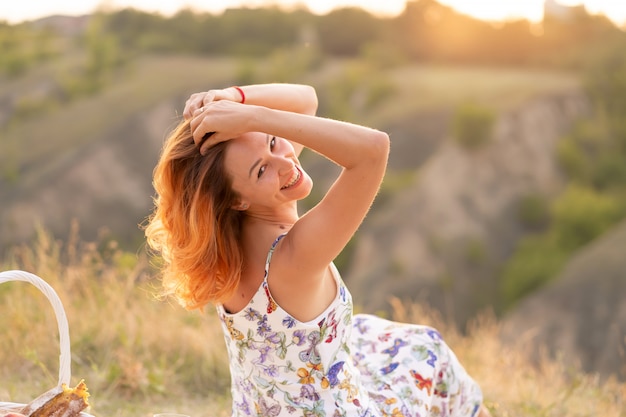 Tender beautiful red-haired girl enjoys the sunset in a field with a hill.