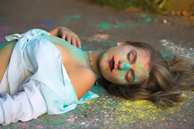 Tender blonde woman with closed eyes covered Holi paint lying on the asphalt