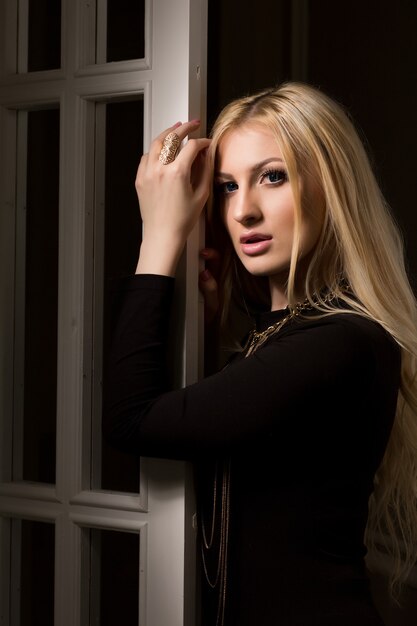 Нежная блондинка с длинными пышными волосами позирует у белой двери с тенью на лице
