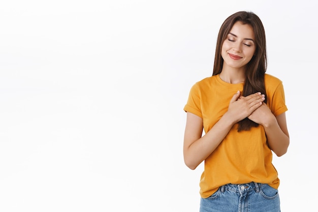 Tender carefree romantic dreamy young brunette woman in yellow tshirt holding hands pressed to heart filled with warmth of love and passion close eyes and smiling recall happy moment