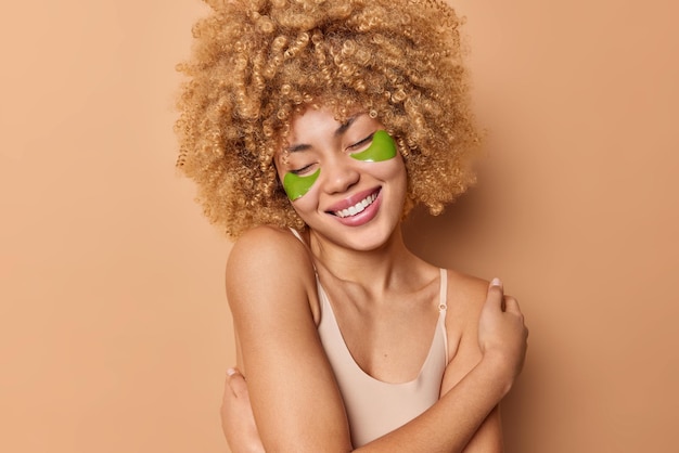 Фото Нежная жизнерадостная европейская женщина с вьющимися густыми волосами обнимает собственное тело, носит футболку, улыбается, нежно наносит гидрогелевые зеленые пятна под глазами, изолированными на коричневом фоне.