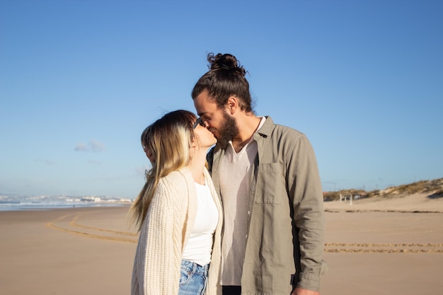 Tender couple walking on beach. Bearded man and woman in glasses looking at each other, kissing. Love, vacation, affection concept