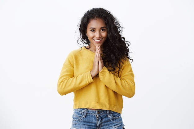 Tender and cute young curly-haired woman in yellow sweater, jeans asking favour, thanking for help, press palms together in pray, smiling joyfully, standing white wall happy