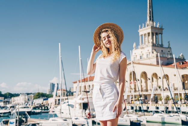 Tender girl in white clothes, covers her eyes from the sun against the background of the sea terminal and ship masts