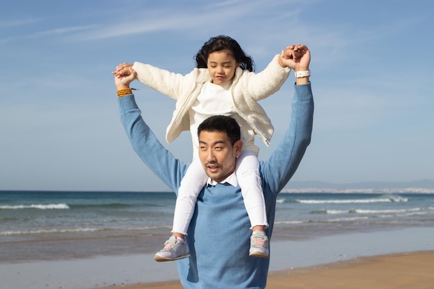 Tender Japanese family walking on beach. Daughter riding on fathers back, looking forward. Leisure, vacation, parenting concept