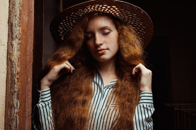 Tender redhead model with closed eyes in straw hat posing in passage. Closeup shot of redhead woman with long hair