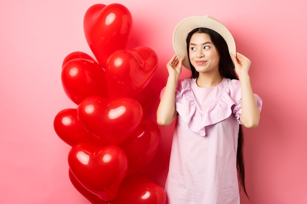 Tenera e romantica ragazza adolescente asiatica che indossa cappello di paglia e vestito alla data, in piedi vicino a palloncini cuore di san valentino e guardando da parte con un sorriso sognante, sfondo rosa.