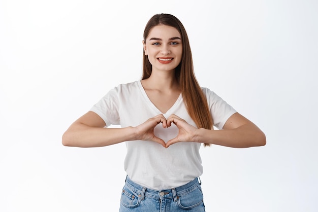 Tender and romantic girl shows heart sign I love you gesture smiling lovely at camera standing in tshirt and jeans against white background Copy space