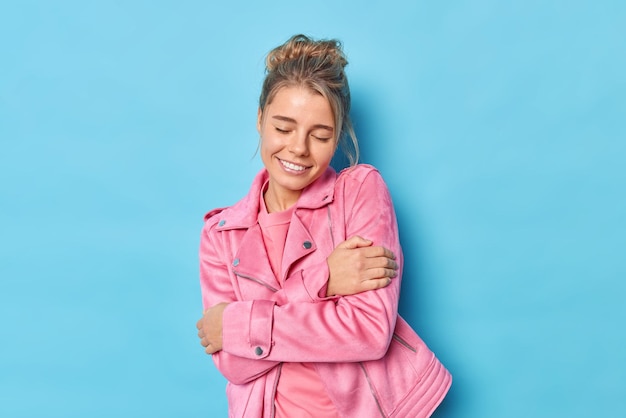 Tender satisfied young lovely woman embraces herself keeps eyes closed dressed in casual pink jacket feels coziness daydreams about something isolated over blue background recalls romantic date