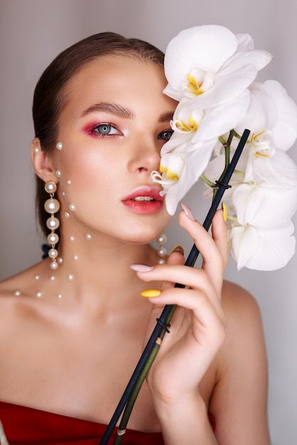 Tender stylish woman with makeup and in pearl earrings covering eye with white orchid flower