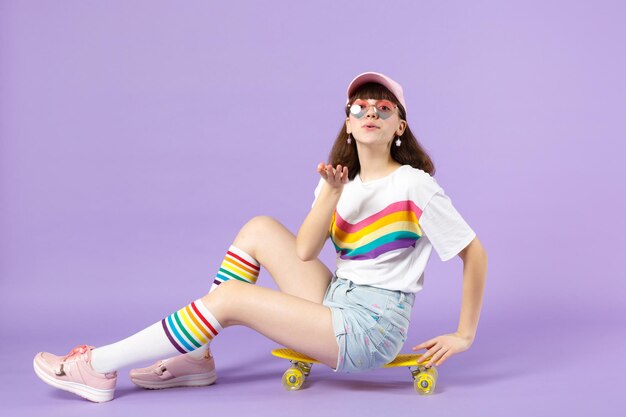 Tender teen girl in vivid clothes, eyeglasses sitting on yellow skateboard blowing sending air kiss isolated on violet pastel background. People sincere emotions lifestyle concept. Mock up copy space.
