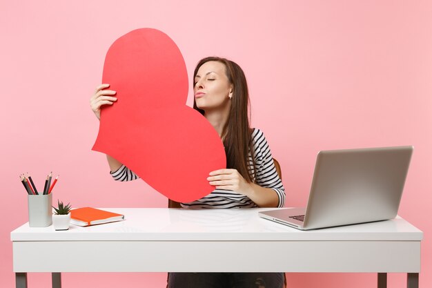 Tender woman blowing lips sending air kiss holding red empty blank heart sit and work at white desk with pc laptop isolated on pastel pink background. Achievement business career concept. Copy space.