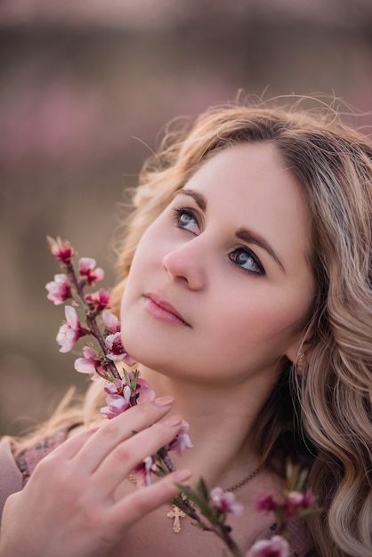 Tender young woman with beautiful curly blonde hair, with clean face, natural makeup, stands in blooming gardens, holds twig with pink inflorescences