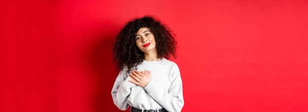 Photo tender young woman with curly hairstyle holding hands on heart and smiling with grateful expression