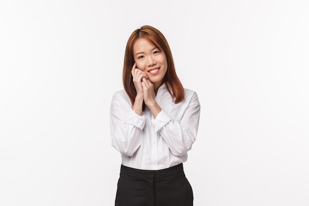 Tenderness, beauty and business concept. Tender charming asian woman, office lady in white shirt and black skirt, softly touching her face and smiling delighted, apply make-up, skincare routine