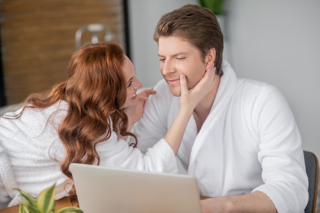 Tenderness. Ginger woman in a white bathrobe and her husband looking happy and in love