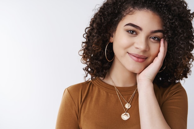 Tenerezza, benessere, concetto di bellezza. bella ragazza bruna dai capelli ricci pura pelle pulita tocco guancia teneramente dolce sorriso fotocamera sentire relax felicità all'interno in piedi sfondo bianco sensualmente