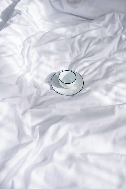 Tenderness. White with black rimmed cup and saucer on clean snow-white crumpled bedding in daylight indoors