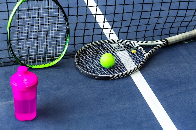 Tennis rackets water bottle and balls against a stretched tennis net on an indoor tennis court Horizontal photo