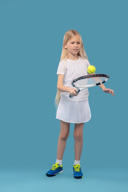 Tennis, warmup. Interested focused longhaired girl in white tshirt and skirt exercising with tennis racket and ball on blue background