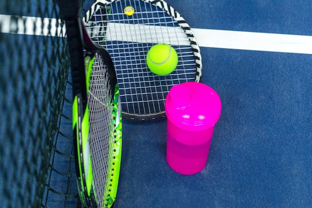 Tennisrackets een fles water en een bal tegen de achtergrond van een uitgerekt tennisnet op een indoor tennisbaan Bovenaanzicht Horizontale foto