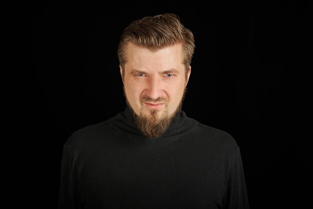 Tense bearded man, black background. Young guy in black polo neck sweater. Suspicious and angry man.