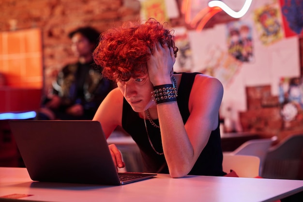 Tense guy with curly hair holding his head in hands and looking at laptop screen