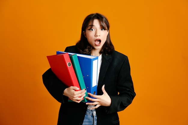 Tense holding folders young beautiful female wearing black jacket isolated on orange background