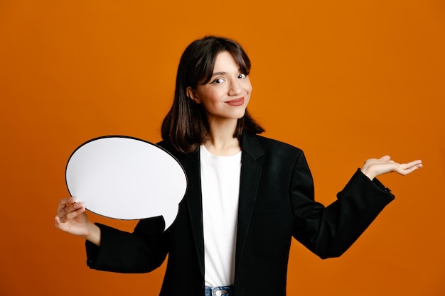 Tense holding speech bubble young beautiful female wearing black jacket isolated on orange background