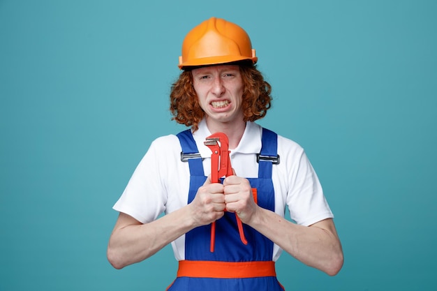 Напряженный молодой строитель в форме держит газовый ключ на синем фоне