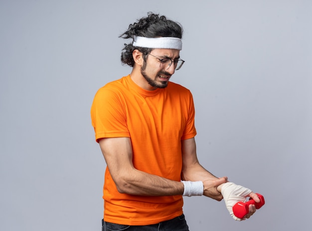 Tense young sporty man wearing headband with wristband with injured wrist wrapped with bandage exercising with dumbbell 