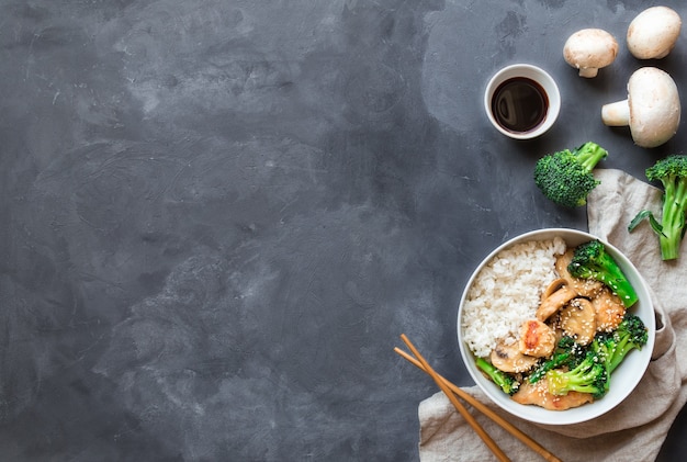 Teriyaki chicken, broccoli and mushrooms stir fry with white rice in bowl on gray concrete background. Asian cuisine. Top view with space for text.