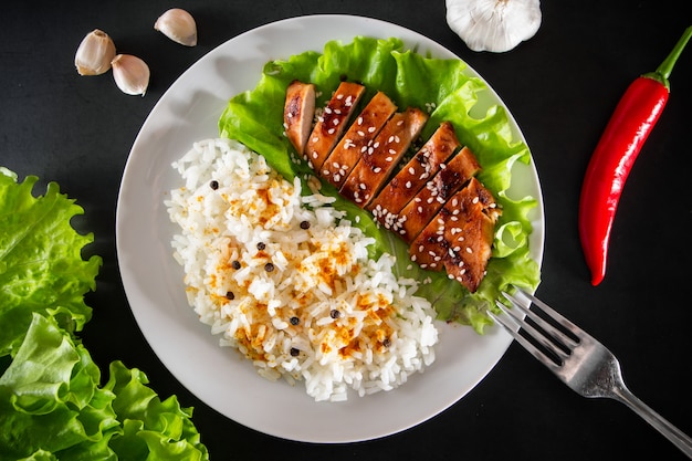 Teriyaki kip met sesamzaadjes, sla en rijst op een witte plaat. Schotel met een vork op een zwarte