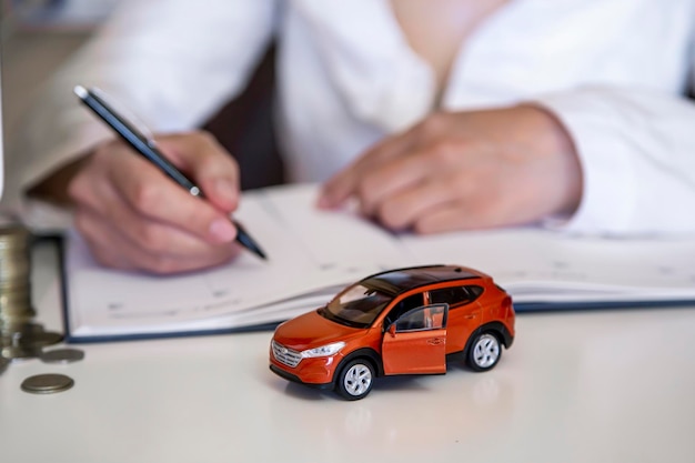 Terracotta color miniature model of a car coins and a hand with a pen on a white table