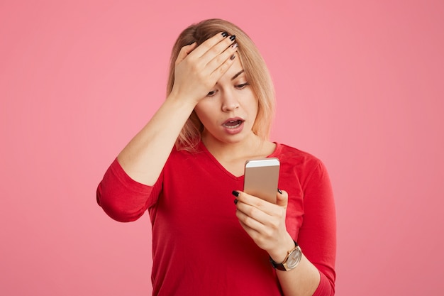 Terrfied blonde young woman stares at camera of mobile phone, keeps hand on forehead, reads bad news on internet website, isolated over pink wall. People, emotions and technology concept