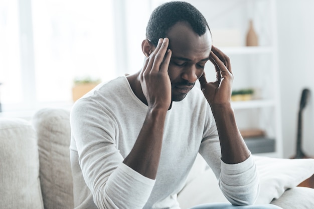 Terrible headache.  Young African man touching head with hands and keeping eyes closed while sitting on the sofa at home