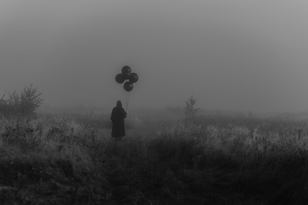 Terrible man in a hooded cloak with balloons in his hand stands in a foggy field. Dark concept of mystical nightmares