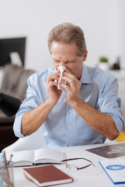 Terrible rhinitis. Disturbed office worker wearing blue shirt closing his eyes while blowing nose